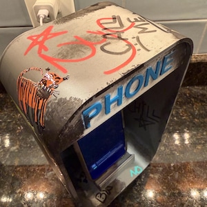 Pode incluir: Um suporte de metal cinzento para telem&oacute;vel com topo curvo, grafiti vermelho com a inscri&ccedil;&atilde;o CALL ME, um desenho de um tigre e a palavra PHONE em letras azuis em relevo. O suporte tem uma ranhura central que segura um smartphone com ecr&atilde; azul.