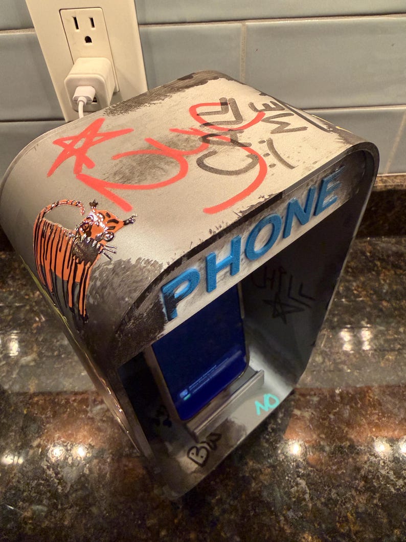 Pode incluir: Um suporte de metal cinzento para telem&oacute;vel com topo curvo, grafiti vermelho com a inscri&ccedil;&atilde;o CALL ME, um desenho de um tigre e a palavra PHONE em letras azuis em relevo. O suporte tem uma ranhura central que segura um smartphone com ecr&atilde; azul.