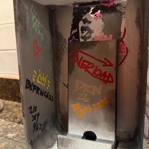 Pode incluir: Um suporte met&aacute;lico para telem&oacute;vel com uma est&eacute;tica industrial desgastada e marcas de estilo graffiti. A superf&iacute;cie apresenta v&aacute;rios elementos desenhados &agrave; m&atilde;o, incluindo o texto "OVERLOAD", "BX", "DXN 26" e "BXPRINCESS", al&eacute;m de n&uacute;meros e um autocolante.