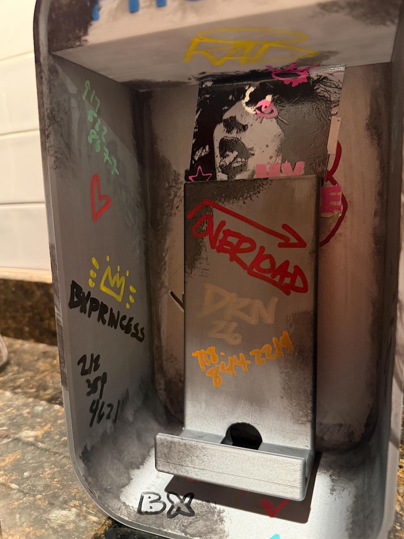 Pode incluir: Um suporte met&aacute;lico para telem&oacute;vel com uma est&eacute;tica industrial desgastada e marcas de estilo graffiti. A superf&iacute;cie apresenta v&aacute;rios elementos desenhados &agrave; m&atilde;o, incluindo o texto "OVERLOAD", "BX", "DXN 26" e "BXPRINCESS", al&eacute;m de n&uacute;meros e um autocolante.