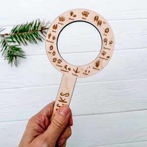 May include: A wooden magnifying glass with a handle and a circular magnifying lens. The lens is surrounded by a ring with various animal tracks and names, including deer, raccoon, badger, bear, elk, skunk, coyote, opossum, turkey, rabbit, fox, squirrel, woodchuck, and beaver.