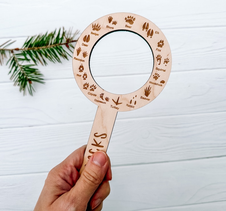 May include: A wooden magnifying glass with a handle and a circular magnifying lens. The lens is surrounded by a ring with various animal tracks and names, including deer, raccoon, badger, bear, elk, skunk, coyote, opossum, turkey, rabbit, fox, squirrel, woodchuck, and beaver.