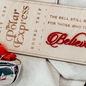 May include: A silver bell with a red ribbon and the word "Believe" engraved on it. A wooden sign with "The Polar Express" and "Believe" in red script is also present. The sign also reads "The bell still rings for those who truly."