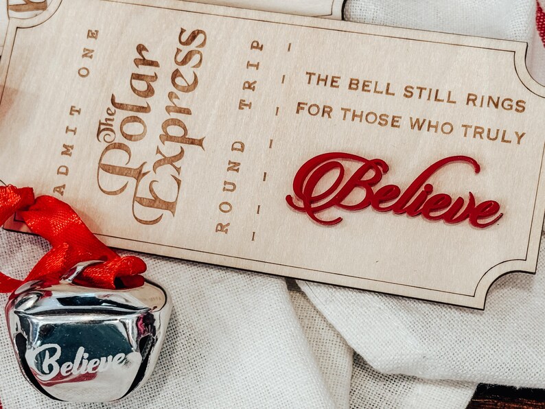 May include: A silver bell with a red ribbon and the word "Believe" engraved on it. A wooden sign with "The Polar Express" and "Believe" in red script is also present. The sign also reads "The bell still rings for those who truly."