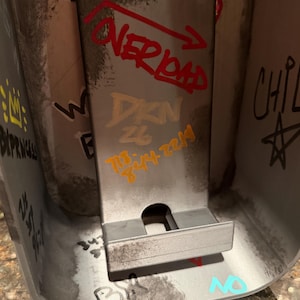 Pode incluir: Um suporte de telefone cinza met&aacute;lico com um painel traseiro retangular e uma pequena borda. A superf&iacute;cie est&aacute; coberta com marcas estilo grafite, incluindo o texto vermelho "OVERLOAD" com uma seta, texto dourado "DKN 26" e n&uacute;meros laranja.