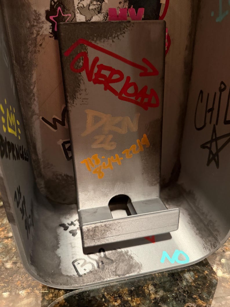 Pode incluir: Um suporte de telefone cinza met&aacute;lico com um painel traseiro retangular e uma pequena borda. A superf&iacute;cie est&aacute; coberta com marcas estilo grafite, incluindo o texto vermelho "OVERLOAD" com uma seta, texto dourado "DKN 26" e n&uacute;meros laranja.