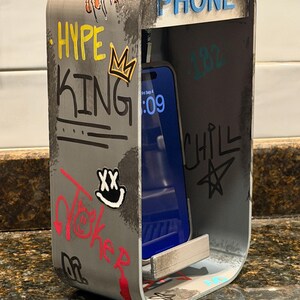 Pode incluir: Um suporte de telefone de metal prateado em forma de cabine telef&oacute;nica com cantos arredondados. O exterior apresenta grafites com as palavras HYPE KING, CHILL e uma cara sorridente. A palavra PHONE est&aacute; no topo. Um smartphone est&aacute; no suporte.