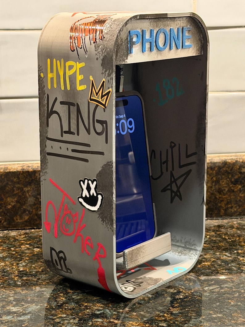 Pode incluir: Um suporte de telefone de metal prateado em forma de cabine telef&oacute;nica com cantos arredondados. O exterior apresenta grafites com as palavras HYPE KING, CHILL e uma cara sorridente. A palavra PHONE est&aacute; no topo. Um smartphone est&aacute; no suporte.