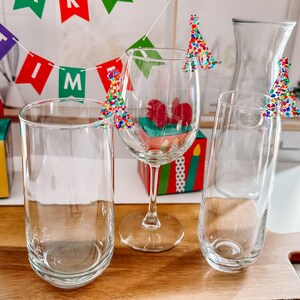 May include: Four clear glass drinking glasses with colorful, confetti-like Christmas tree toppers. The glasses are on a wooden surface.