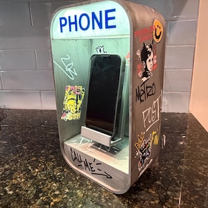 May include: A gray, rectangular phone stand designed like a miniature telephone booth with an illuminated top sign reading PHONE. The exterior features graffiti-style stickers and text, including CALL ME, while a smartphone rests in the center.