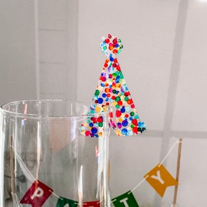 May include: A clear glass with a colorful confetti party hat topper. The hat is made of clear plastic with a confetti pattern in shades of red, blue, green, yellow, and orange.