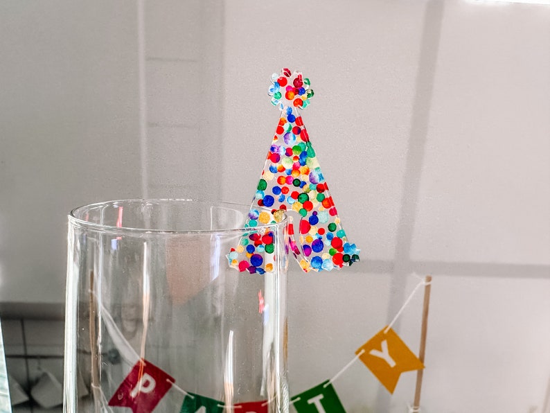 May include: A clear glass with a colorful confetti party hat topper. The hat is made of clear plastic with a confetti pattern in shades of red, blue, green, yellow, and orange.