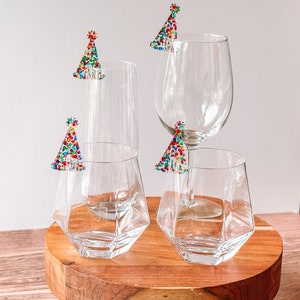 May include: Four clear glass drinking glasses with colorful confetti party hats on the rims. The glasses are on a wooden coaster.