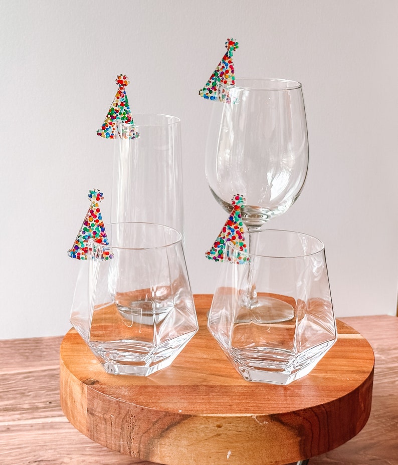 May include: Four clear glass drinking glasses with colorful confetti party hats on the rims. The glasses are on a wooden coaster.