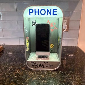 May include: A rectangular phone charging station designed to resemble a vintage telephone booth. The light green structure features the word PHONE in blue text at the top and CALL ME written in black marker at the base, with various graffiti-style drawings.