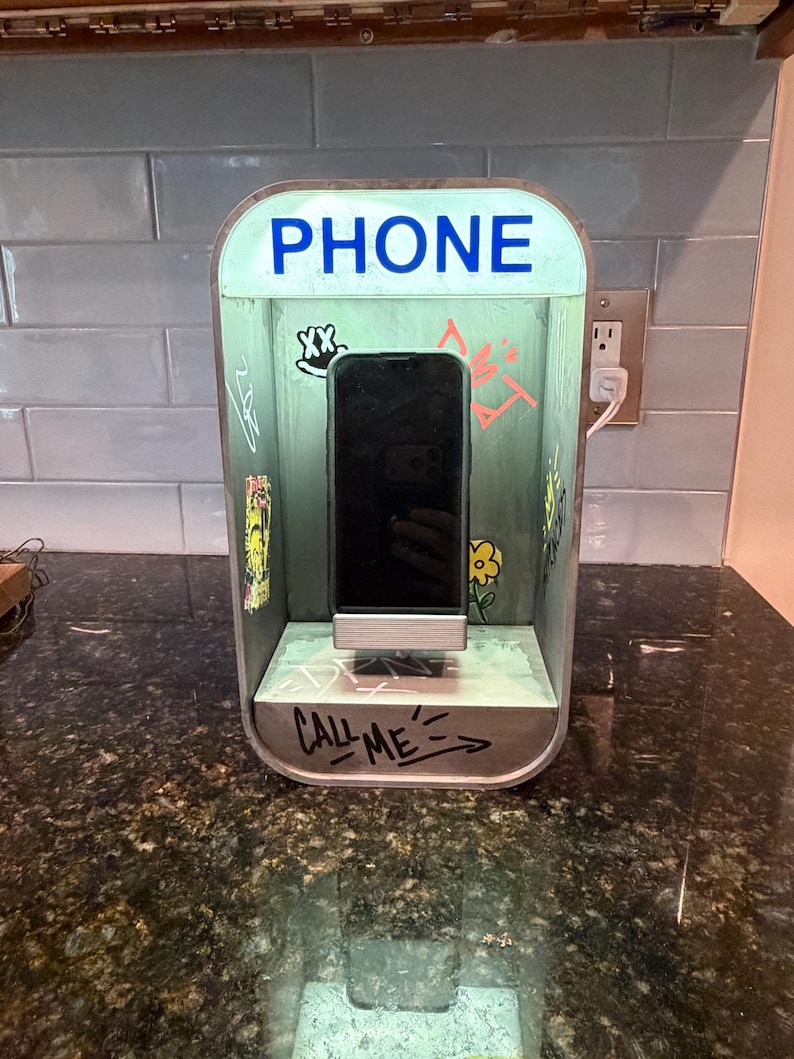May include: A rectangular phone charging station designed to resemble a vintage telephone booth. The light green structure features the word PHONE in blue text at the top and CALL ME written in black marker at the base, with various graffiti-style drawings.