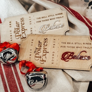 May include: Two wooden Polar Express tickets with the words "Believe" and two silver bells with red ribbons. The tickets have the text "The Bell Still Rings For Those Who Truly". The bells have the word "Believe" on them.