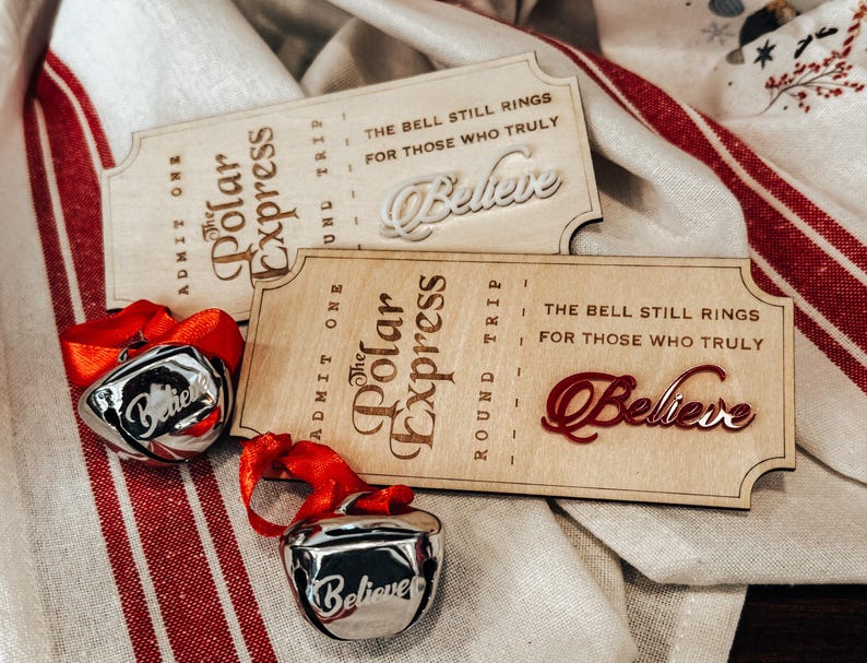 May include: Two wooden Polar Express tickets with the words "Believe" and two silver bells with red ribbons. The tickets have the text "The Bell Still Rings For Those Who Truly". The bells have the word "Believe" on them.