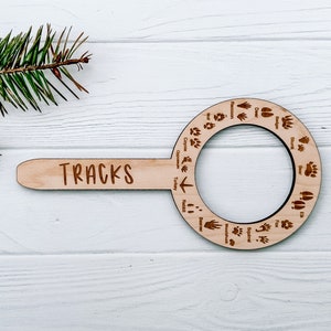 Könnte beinhalten: Ein Holzwerkzeug zur Identifizierung von Spuren mit dem Wort "TRACKS" auf dem Griff. Das Werkzeug hat einen kreisförmigen Ausschnitt mit verschiedenen Tierpfotenabdrücken, die um den Rand herum gedruckt sind.