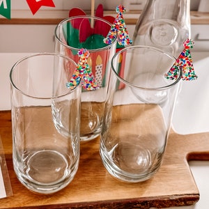 May include: Three clear glass tumblers with colorful confetti-like Christmas tree decorations on the rims. The glasses are sitting on a wooden surface.