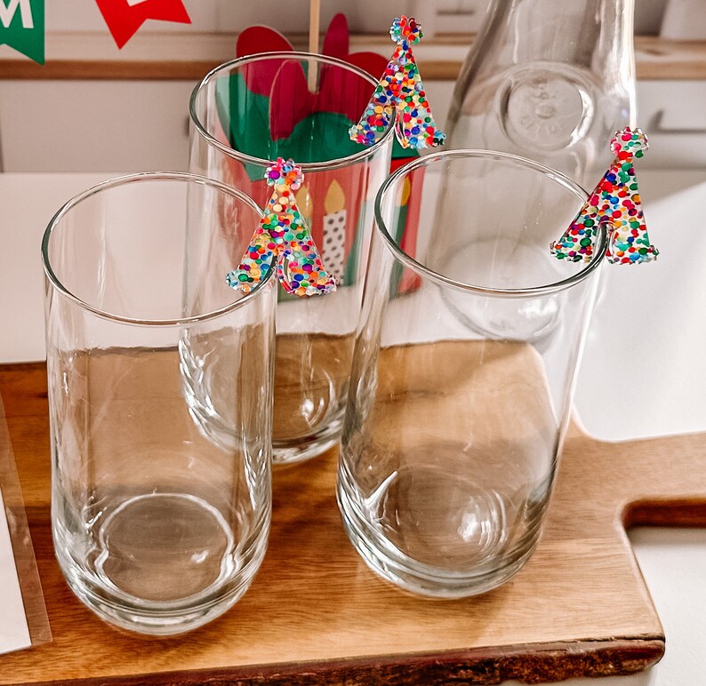 May include: Three clear glass tumblers with colorful confetti-like Christmas tree decorations on the rims. The glasses are sitting on a wooden surface.