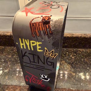 Pode incluir: Um suporte de metal cinzento curvo com arte estilo graffiti, incluindo um tigre laranja, uma coroa amarela e o texto HYPE KING em amarelo e preto. Outras marcas incluem uma estrela vermelha, a palavra JOKE em vermelho e um rosto sorridente branco com olhos XX.