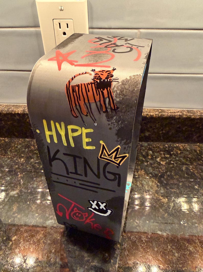 Pode incluir: Um suporte de metal cinzento curvo com arte estilo graffiti, incluindo um tigre laranja, uma coroa amarela e o texto HYPE KING em amarelo e preto. Outras marcas incluem uma estrela vermelha, a palavra JOKE em vermelho e um rosto sorridente branco com olhos XX.