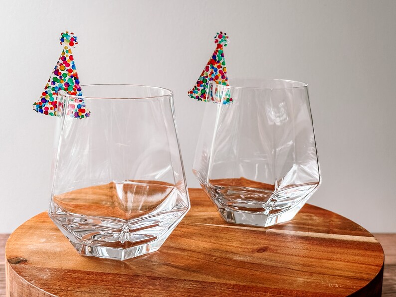 May include: Two clear glass tumblers with a geometric design, each with a colorful confetti party hat sticker on the rim.