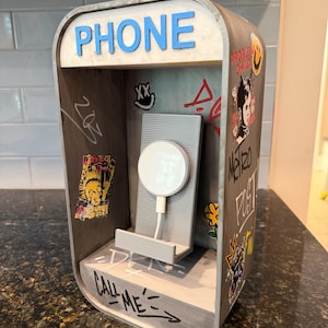 May include: A gray 3D-printed phone booth charging stand featuring the word PHONE in blue letters at the top. The interior walls are decorated with various stickers and graffiti-style markings, including the text CALL ME at the base.