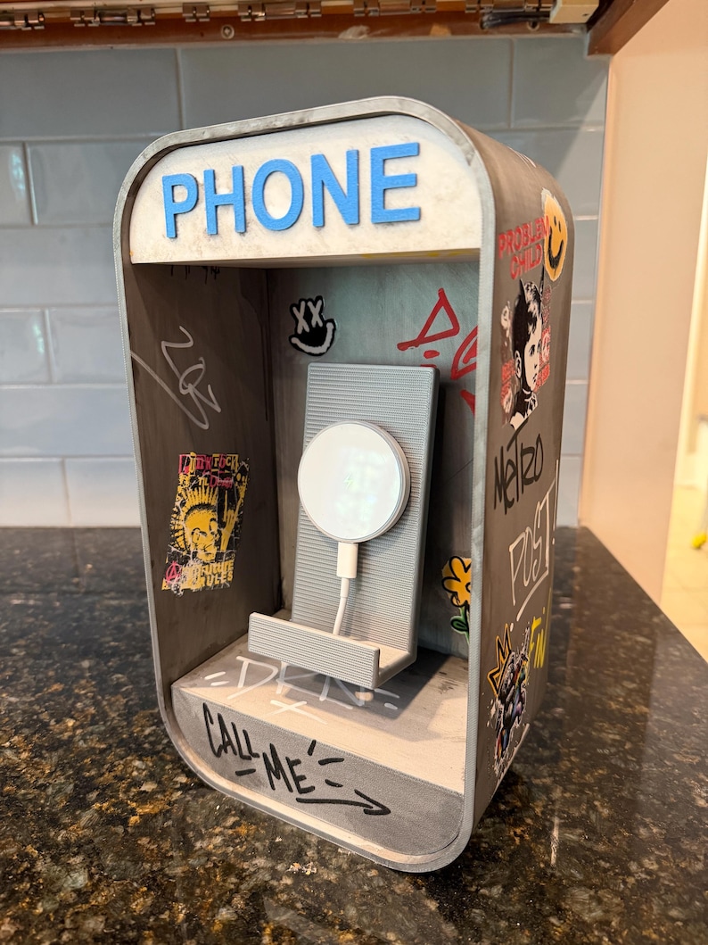 May include: A gray 3D-printed phone booth charging stand featuring the word PHONE in blue letters at the top. The interior walls are decorated with various stickers and graffiti-style markings, including the text CALL ME at the base.