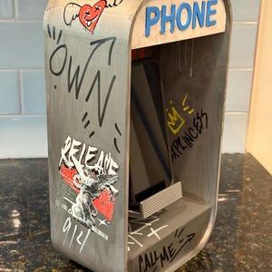 May include: A gray wooden phone stand shaped like a vintage booth, featuring graffiti-style graphics, a red heart with wings, and the text "PHONE", "OWN", "RELEASE", "XPRINCESS", and "CALL ME". A smartphone rests inside the stand on a small ledge.