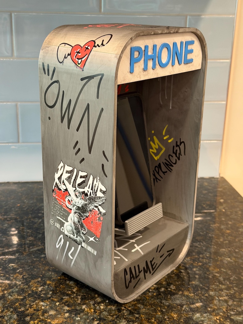 May include: A gray wooden phone stand shaped like a vintage booth, featuring graffiti-style graphics, a red heart with wings, and the text "PHONE", "OWN", "RELEASE", "XPRINCESS", and "CALL ME". A smartphone rests inside the stand on a small ledge.