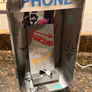 Pode incluir: Um suporte de carregamento de telefone cinza projetado como uma cabine telef&ocirc;nica em miniatura com a palavra PHONE em letras azuis no topo. O interior apresenta texto estilo grafite, incluindo OVERLOAD, DKN e CHILL, com um cabo USB branco.