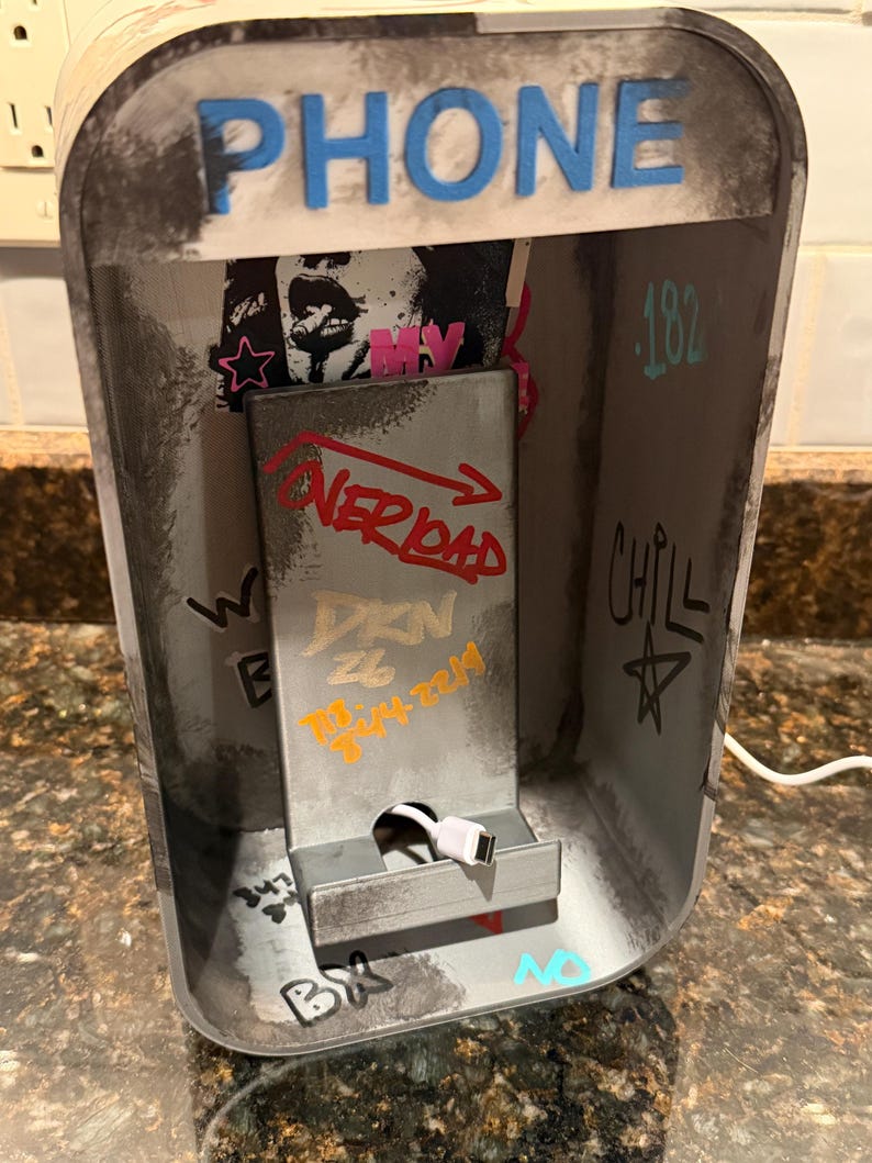 Pode incluir: Um suporte de carregamento de telefone cinza projetado como uma cabine telef&ocirc;nica em miniatura com a palavra PHONE em letras azuis no topo. O interior apresenta texto estilo grafite, incluindo OVERLOAD, DKN e CHILL, com um cabo USB branco.