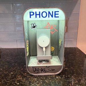 May include: A phone charging station shaped like a miniature telephone booth with a light-up sign reading PHONE. The interior features graffiti-style drawings, a yellow flower, and the text CALL ME. A white circular magnetic charger is mounted inside.