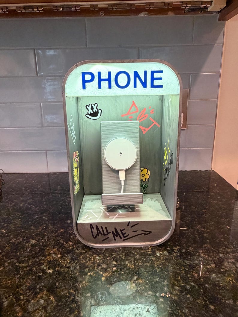 May include: A phone charging station shaped like a miniature telephone booth with a light-up sign reading PHONE. The interior features graffiti-style drawings, a yellow flower, and the text CALL ME. A white circular magnetic charger is mounted inside.