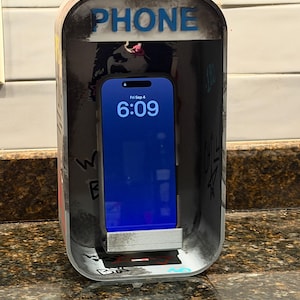 Pode incluir: Um suporte retangular para telem&oacute;vel concebido como uma cabine telef&oacute;nica em miniatura com um exterior desgastado e coberto de graffiti. O topo apresenta a palavra PHONE em letras azuis. Um smartphone est&aacute; encaixado, exibindo as 6:09.