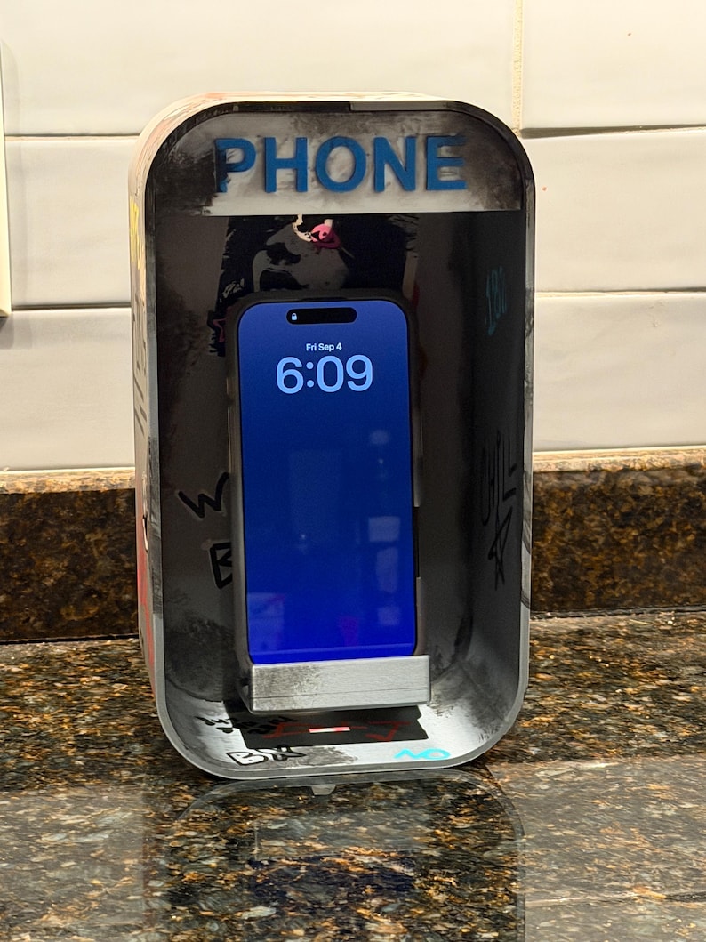 Pode incluir: Um suporte retangular para telem&oacute;vel concebido como uma cabine telef&oacute;nica em miniatura com um exterior desgastado e coberto de graffiti. O topo apresenta a palavra PHONE em letras azuis. Um smartphone est&aacute; encaixado, exibindo as 6:09.