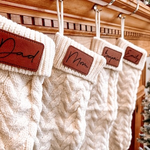 Può includere: Quattro calze di Natale in maglia bianca con etichette in pelle marrone. Le calze sono appese a un camino in legno e i nomi sulle etichette sono Papà, Mamma, Benjamin ed Emily.