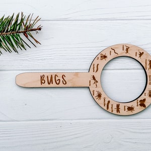 May include: A wooden bug identification tool with a magnifying glass and a handle that says "BUGS". The tool features illustrations of different bugs, including a spider, caterpillar, dragonfly, and beetle.
