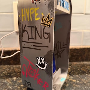 Pode incluir: Um suporte de telefone cinzento, retangular e de estrutura aberta com gr&aacute;ficos estilo graffiti, incluindo o texto HYPE KING, PHONE e um &iacute;cone de coroa. A superf&iacute;cie apresenta v&aacute;rios autocolantes e marcas pretas, amarelas, vermelhas e brancas.