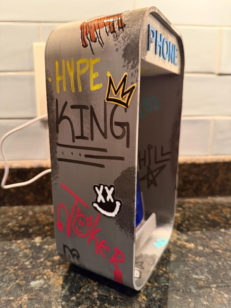 Pode incluir: Um suporte de telefone cinzento, retangular e de estrutura aberta com gr&aacute;ficos estilo graffiti, incluindo o texto HYPE KING, PHONE e um &iacute;cone de coroa. A superf&iacute;cie apresenta v&aacute;rios autocolantes e marcas pretas, amarelas, vermelhas e brancas.