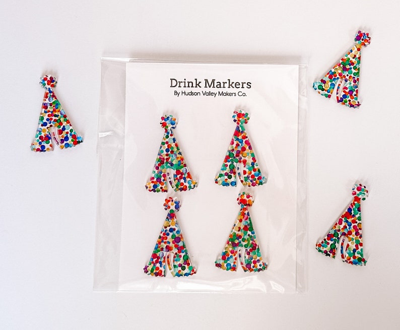 May include: Set of six colorful party hat drink markers. The markers are made of white acrylic and feature a confetti pattern. The markers are packaged in a clear plastic bag with a white card that reads "Drink Markers By Hudson Valley Makers Co."