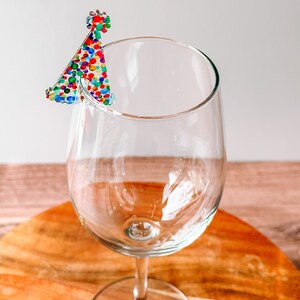 May include: A clear glass wine glass with a colorful party hat charm attached to the rim.