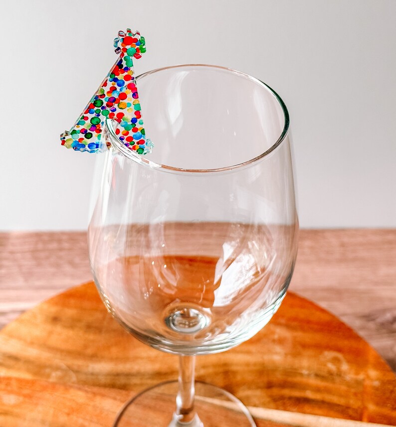 May include: A clear glass wine glass with a colorful party hat charm attached to the rim.