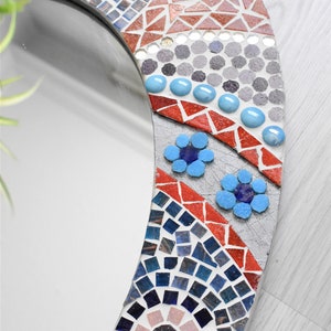 May include: A round mirror with a mosaic frame. The frame is made of small, colorful tiles in shades of blue, red, and gray. The mirror is surrounded by a decorative border of flowers and swirls.