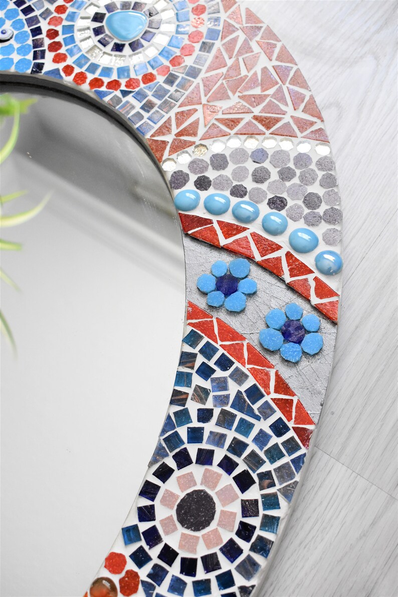 May include: A round mirror with a mosaic frame. The frame is made of small, colorful tiles in shades of blue, red, and gray. The mirror is surrounded by a decorative border of flowers and swirls.