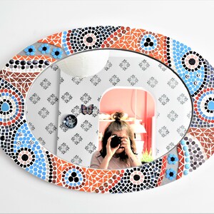 May include: A round mirror with a mosaic frame. The frame is made of small, colorful tiles in shades of blue, red, and white. The mirror is oval-shaped and has a reflective surface.