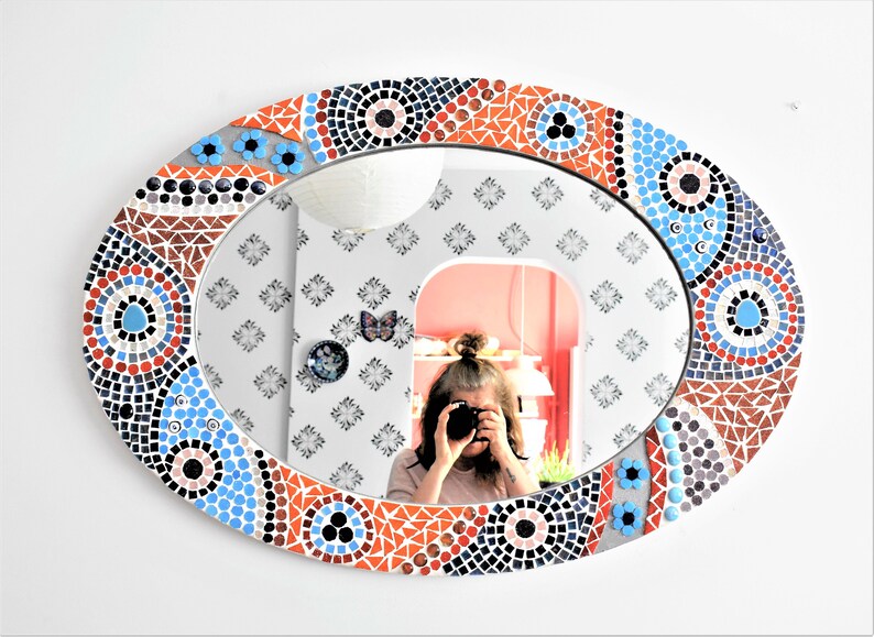 May include: A round mirror with a mosaic frame. The frame is made of small, colorful tiles in shades of blue, red, and white. The mirror is oval-shaped and has a reflective surface.