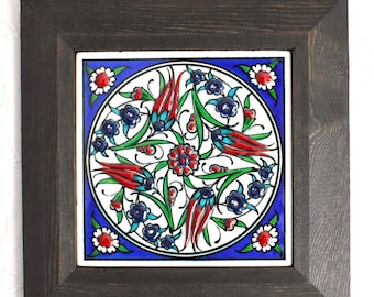 May include: A framed ceramic tile with a blue, white, red, and green floral design. The tile is surrounded by a dark wood frame.